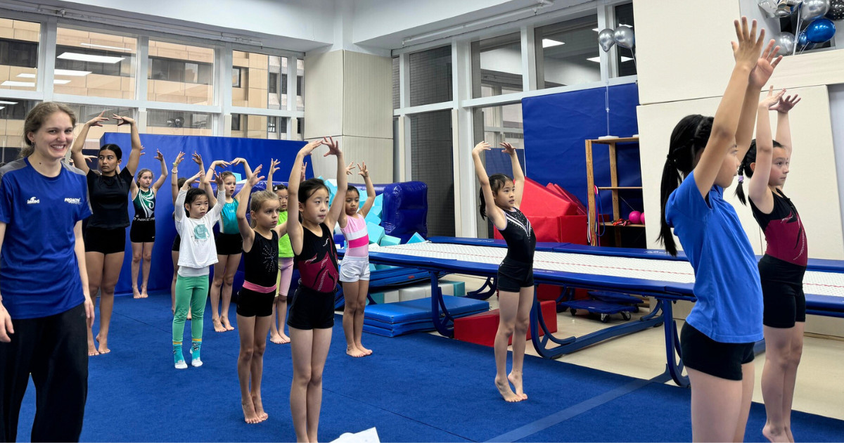 A smiling young girl in a blue leotard jumping on a trampoline during a holiday sports camp.