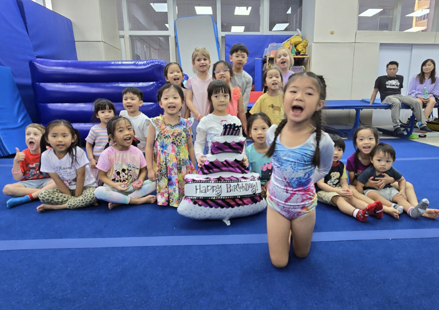 A large birthday cake on a table surrounded by children in a decorated gymnastics gym.