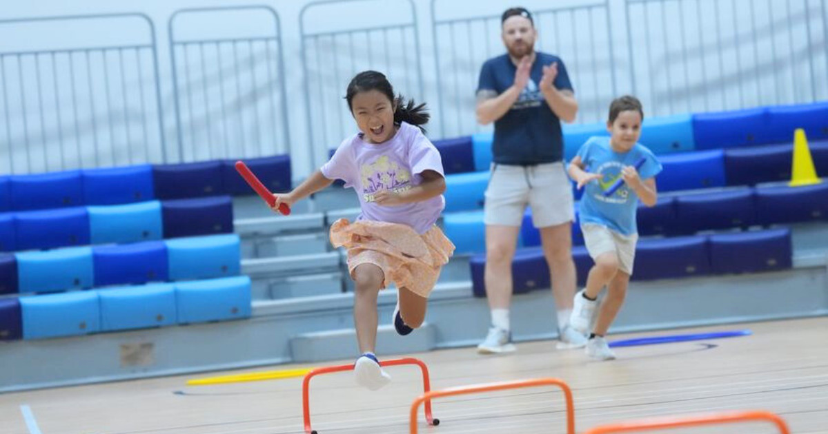 Children playing a variety of games including tennis and gymnastics in a large indoor hall.