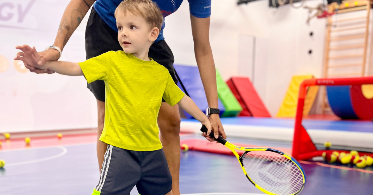 Small child gripping a mini tennis racket and hitting a low-compression yellow ball over a junior net.