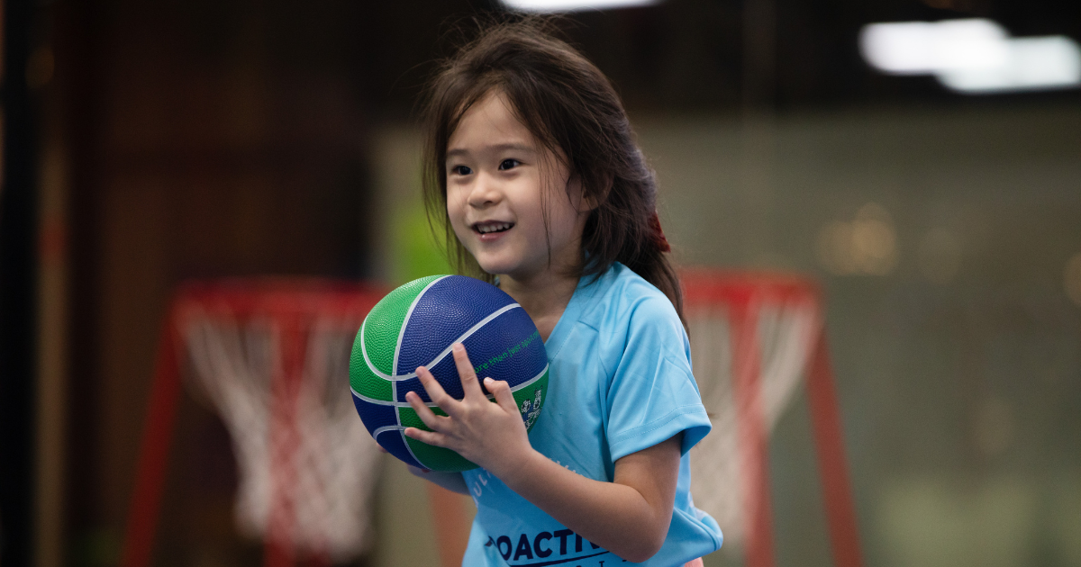 Diverse group of children holding various sports equipment including a soccer ball, basketball, and tennis racket.
